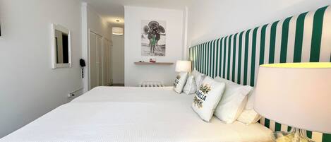 Habitación de hotel con una cama grande, cabecera a rayas, mesita de noche con lámpara y estante en la pared con almohadones decorativos.