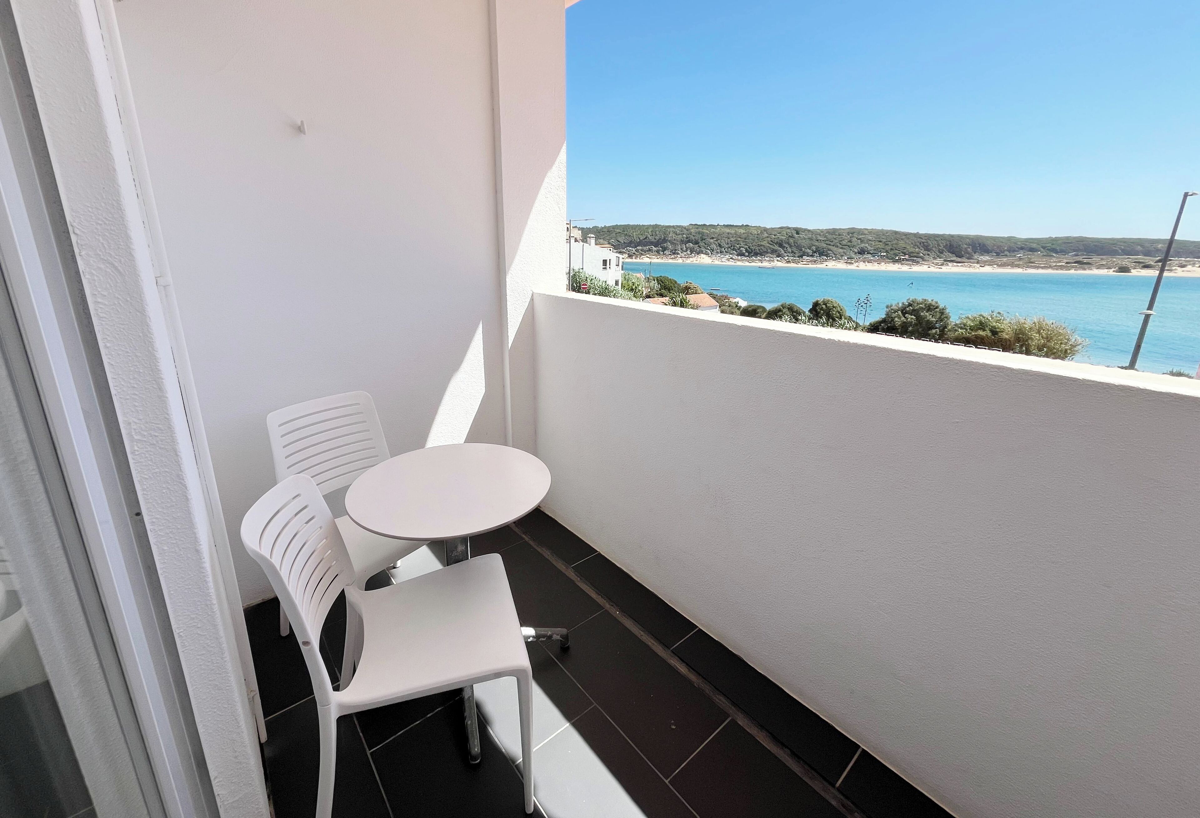 A balcony with a white table and chairs, overlooking a beach with clear blue water and a hilly landscape.