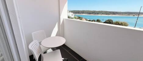 A balcony with a white table and chairs, overlooking a beach with clear blue water and a hilly landscape.