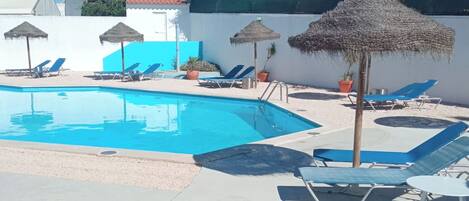 A swimming pool with lounge chairs and thatched umbrellas, surrounded by a white wall and a building with a dome and spires in the background.