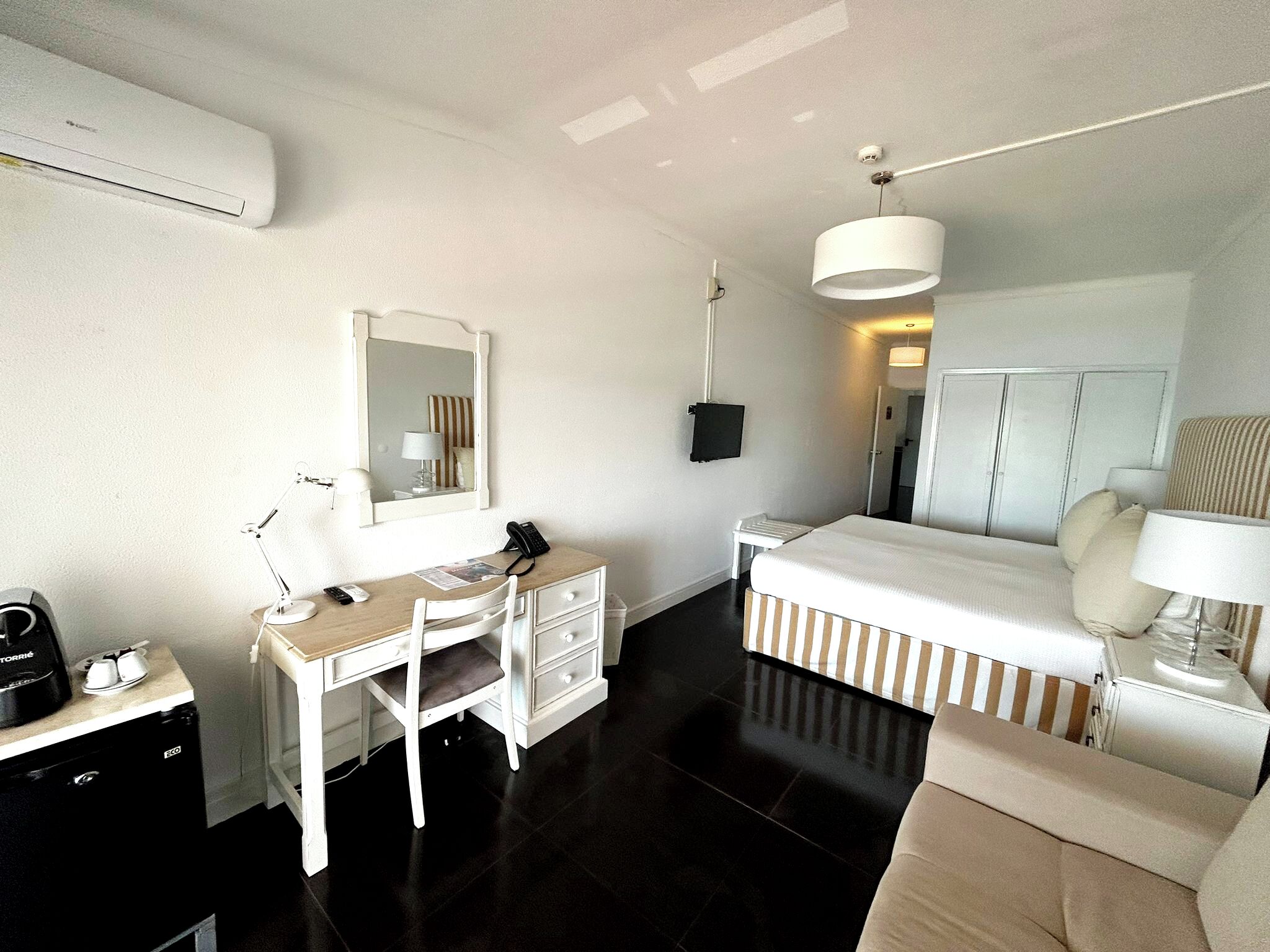 A hotel room with a bed, a desk with a chair, a mirror, a bedside table, a lamp, and an air conditioning unit.