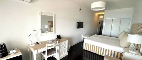 A hotel room with a bed, a desk with a chair, a mirror, a bedside table, a lamp, and an air conditioning unit.