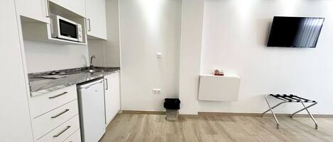 A modern kitchen with white cabinets, a microwave, and a built-in oven. There is a small shelf with a TV mounted on the wall and a chair in the corner.