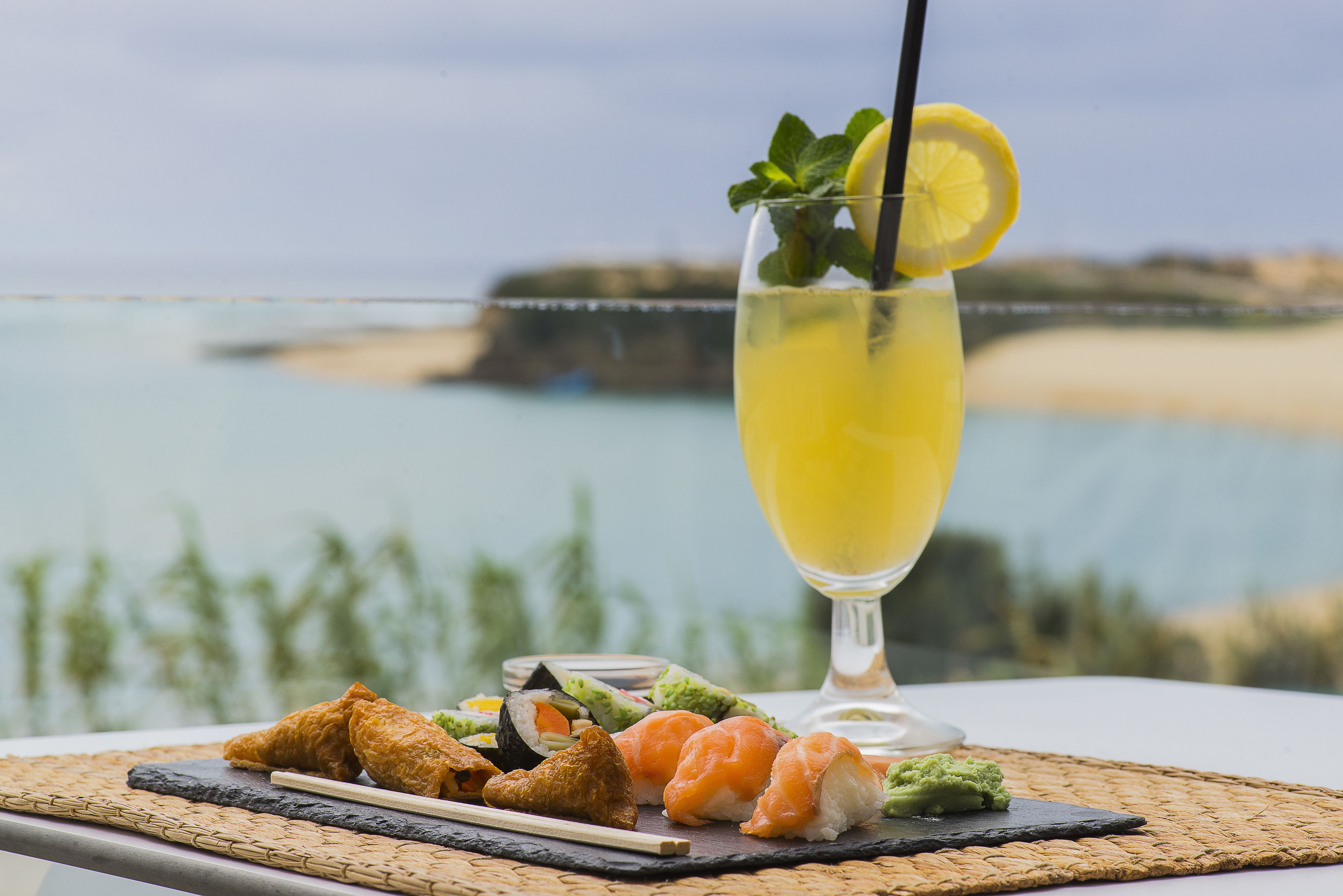 A table with sushi, a glass of yellow drink, and a view of a beach.