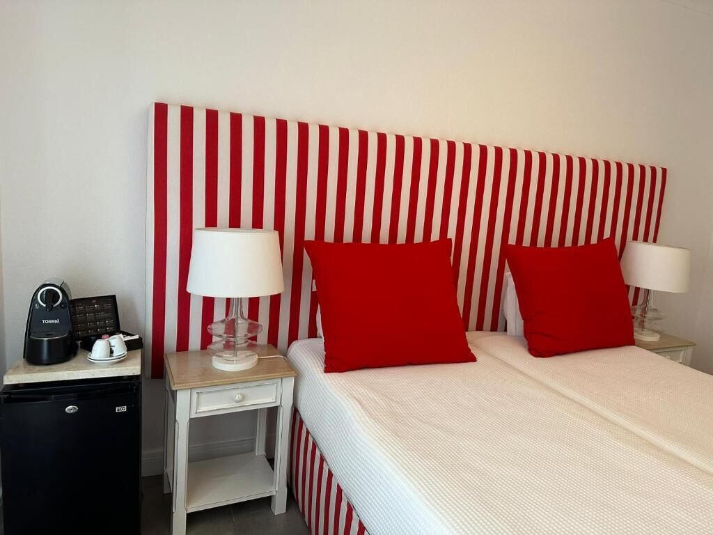 A hotel room with a striped headboard, two red pillows, a bed with a white cover, a nightstand with a lamp, and a small black refrigerator.