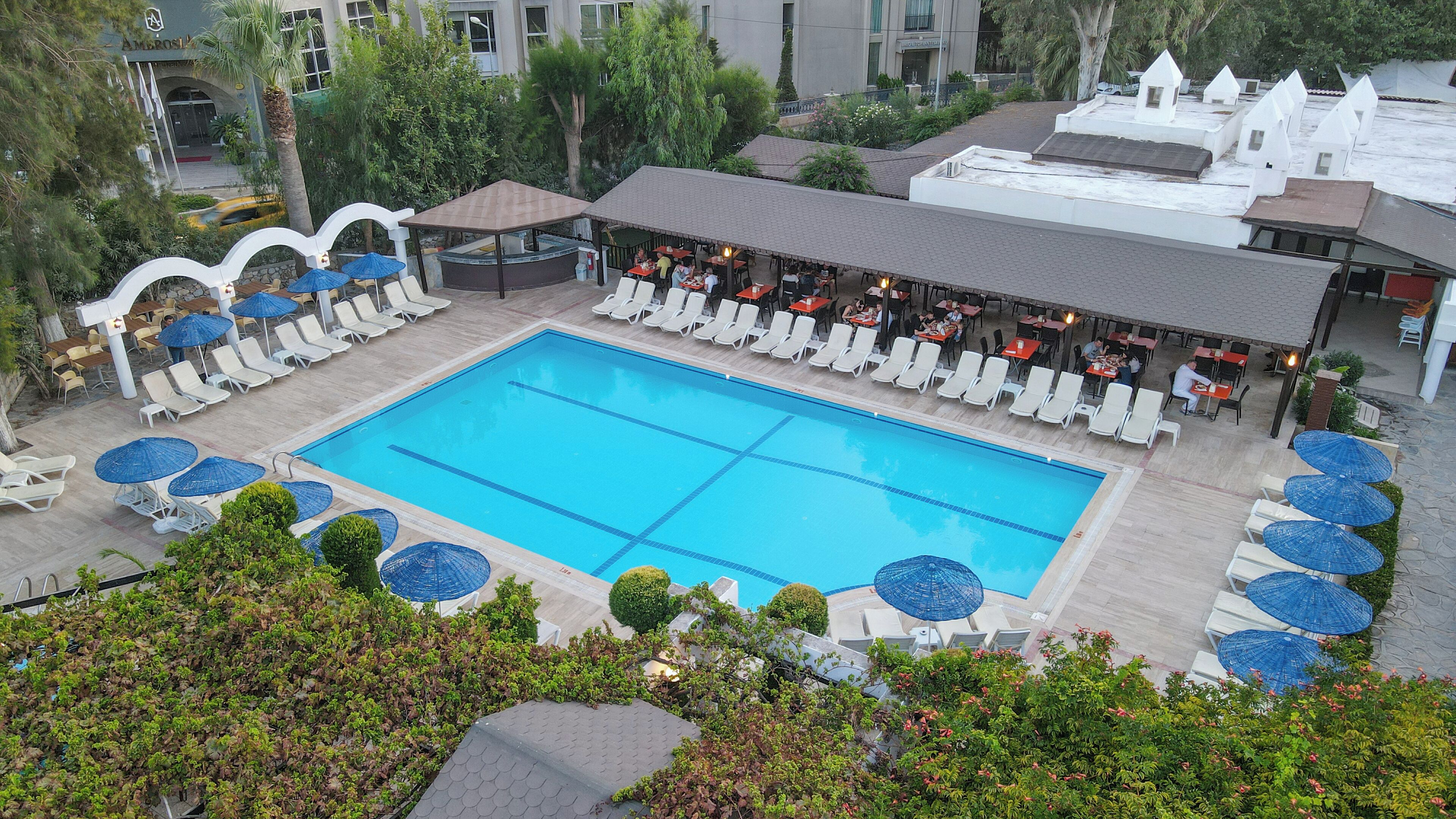 A large swimming pool surrounded by sun loungers and umbrellas, with a covered seating area and a view of the surrounding buildings.