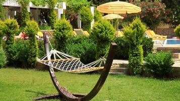 A hammock chair in a lush garden with a pool and sun loungers in the background.