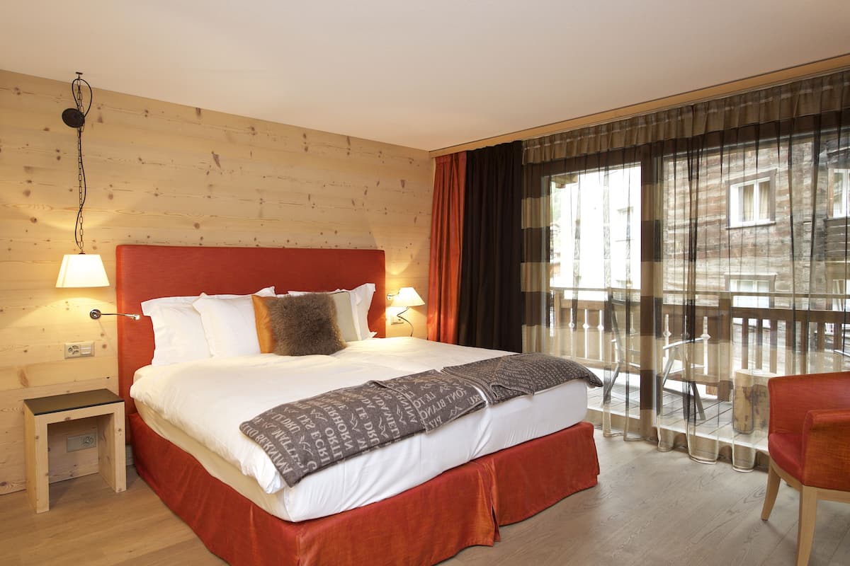 Habitación de hotel con pared de madera, una cama grande con cabecera roja, dos lámparas de noche, una ventana con cortinas y un balcón con una silla.