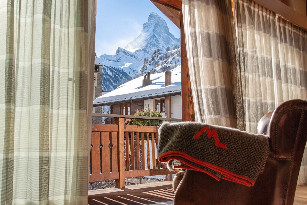 Vista desde una habitación con balcón de madera, con vistas a una montaña y su cima nevada.