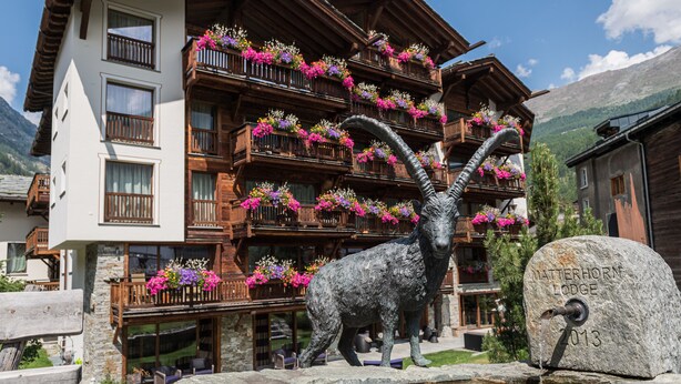 A mountain lodge with multiple balconies adorned with colorful flowers, a stone statue of a goat, and a sign indicating the lodge's name and number.
