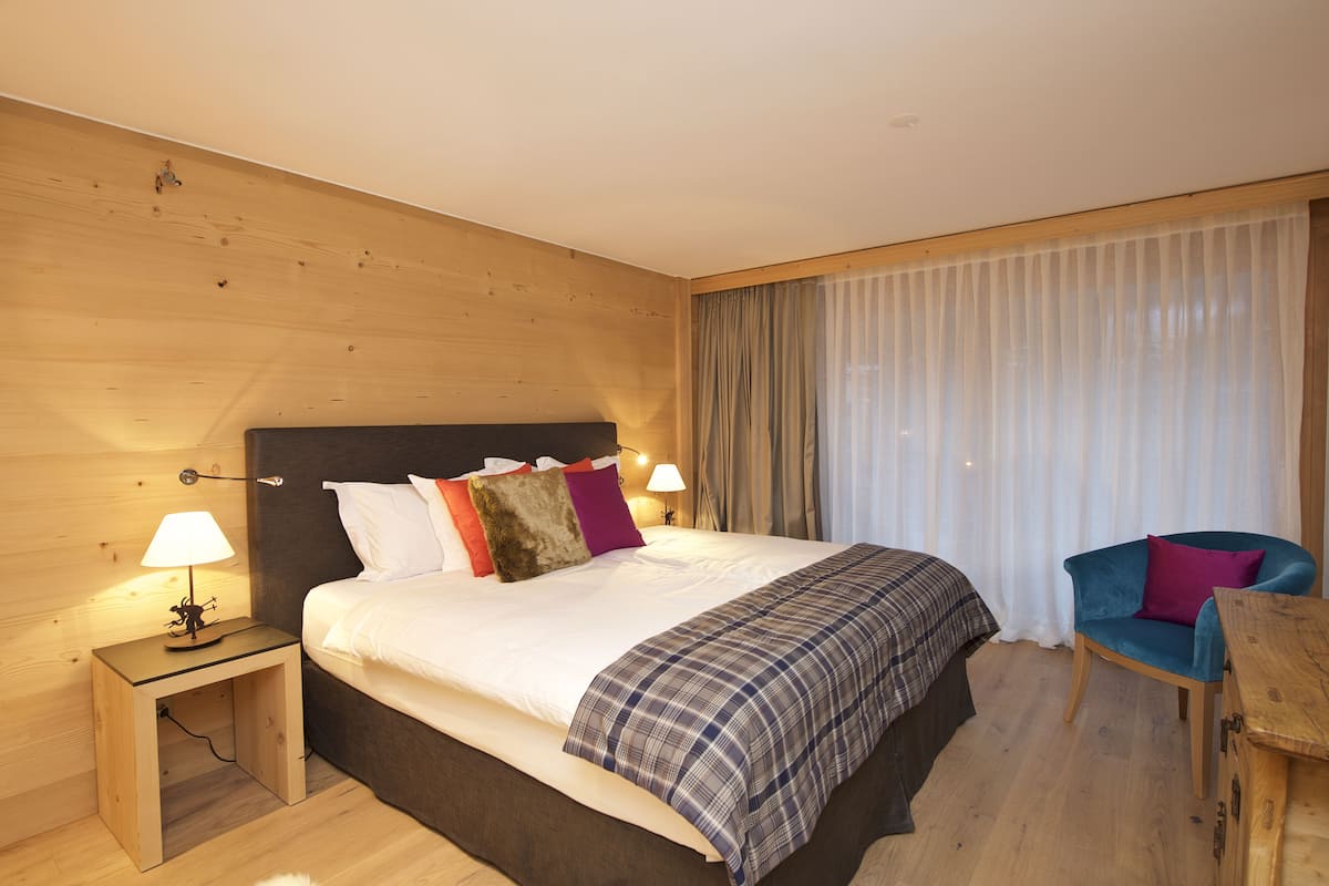 Una habitación de hotel con cabecera de madera, una cama con muchos almohadones, una mesita de noche con lámpara, una silla y una mesa de madera.