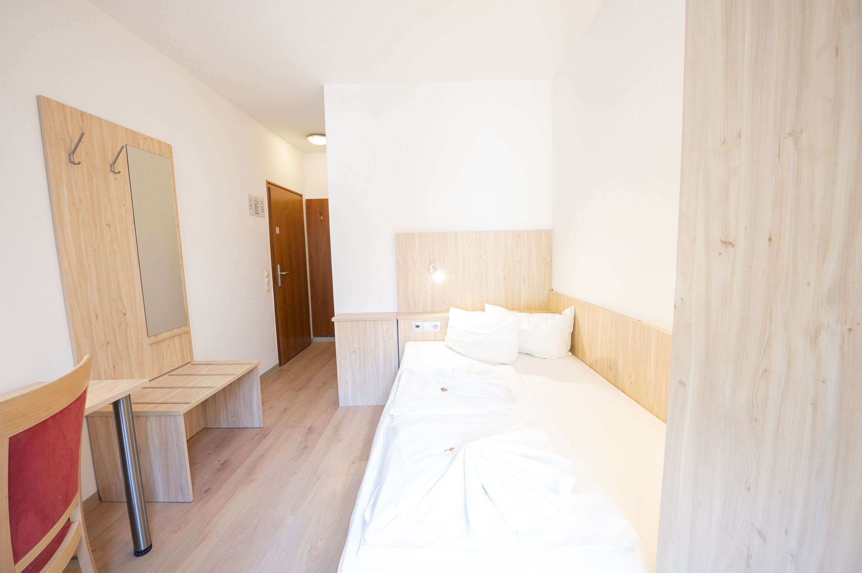 A hotel room with a wooden headboard, a bed with white linens, a wooden bench, a small table, and a mirror.
