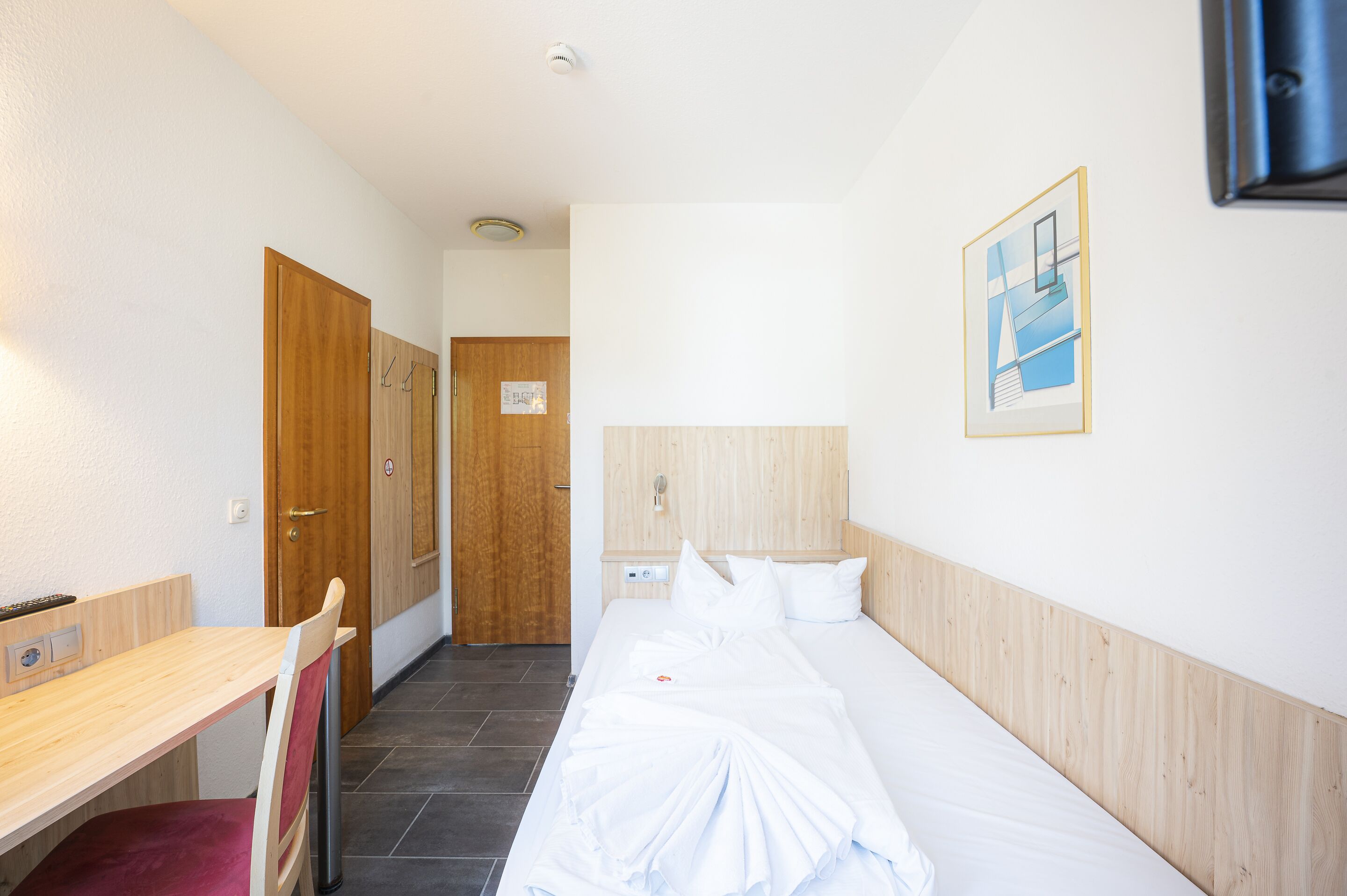 Habitación de hotel con cabecera de madera, cama con ropa blanca, escritorio con silla y puerta de madera.
