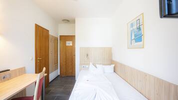 Habitación de hotel con cabecera de madera, cama con ropa blanca, escritorio con silla y puerta de madera.