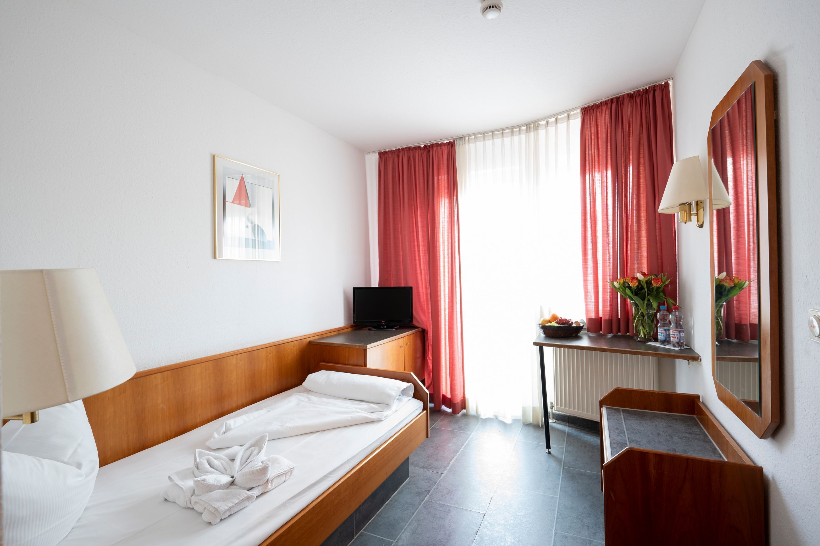 A hotel room with a single bed, a desk with a television, a window with red curtains, a mirror, and a small table with a vase of flowers.