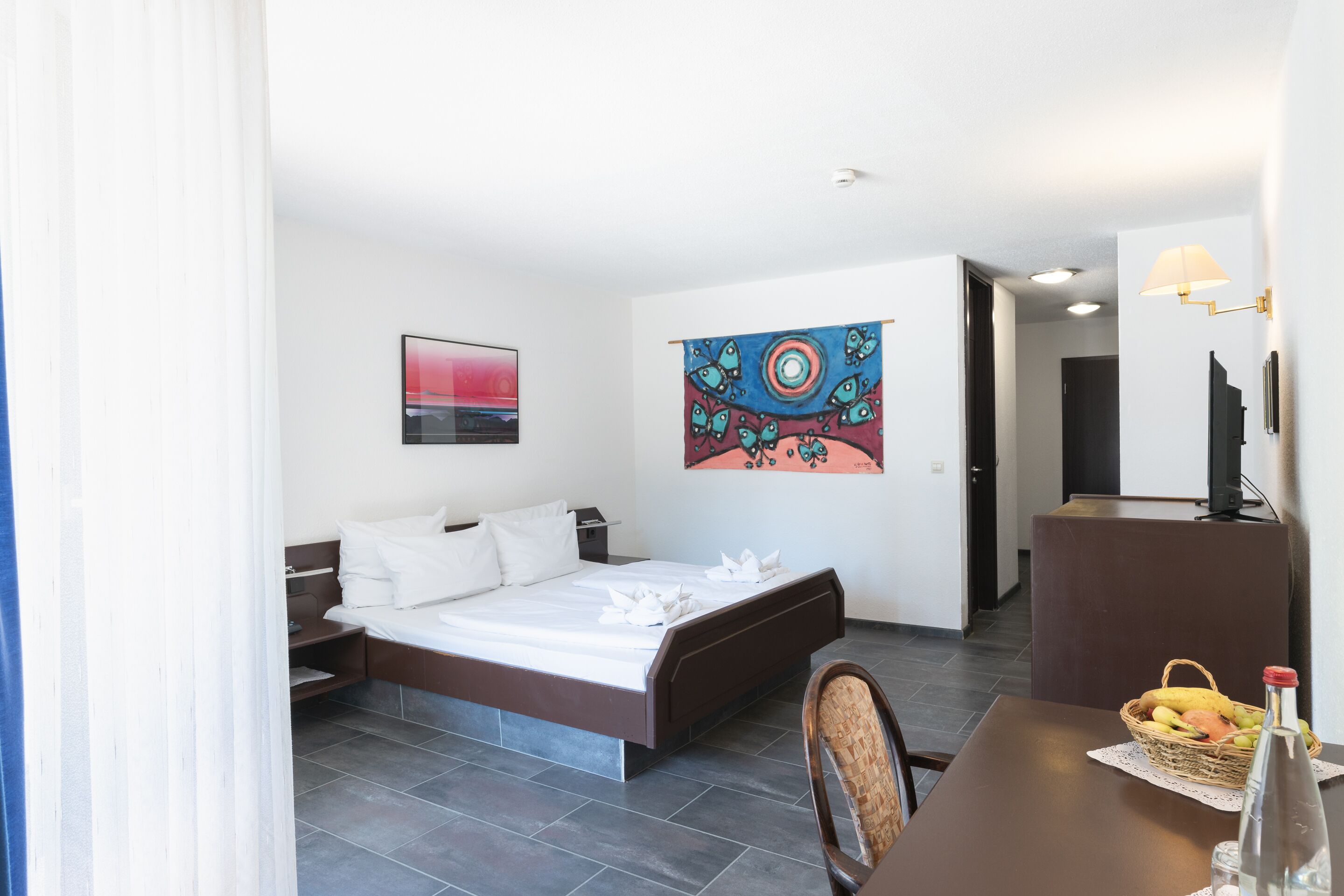 Una habitación de hotel con una cama, un escritorio con una silla, una mesa de comedor y una cesta de fruta, y un tapiz en la pared.
