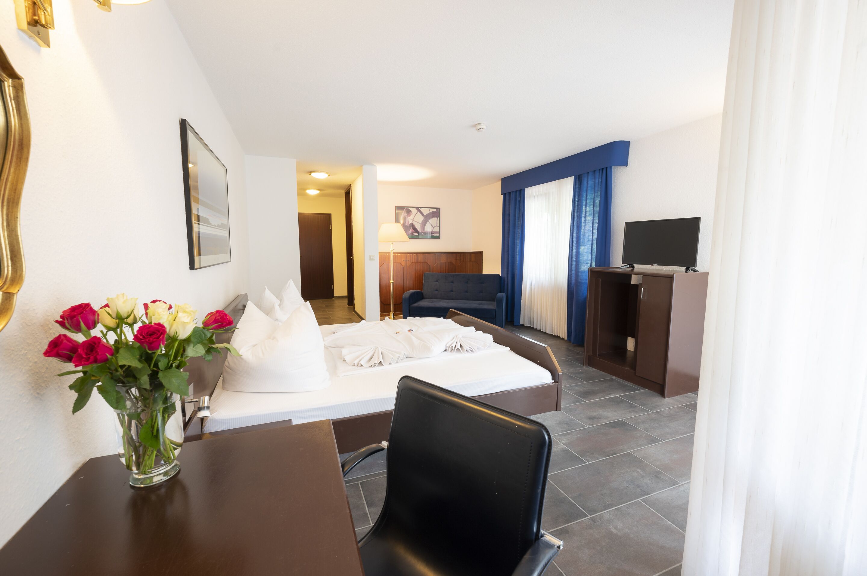 Habitación de hotel con cama, escritorio, silla, televisión, lámpara de escritorio y un florero con flores.