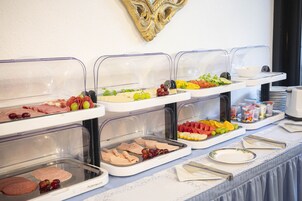 En buffet med forskellige spiser, herunder kød, oste, frugt og grøntsager, anrettet på et bord med et blonde-dug.