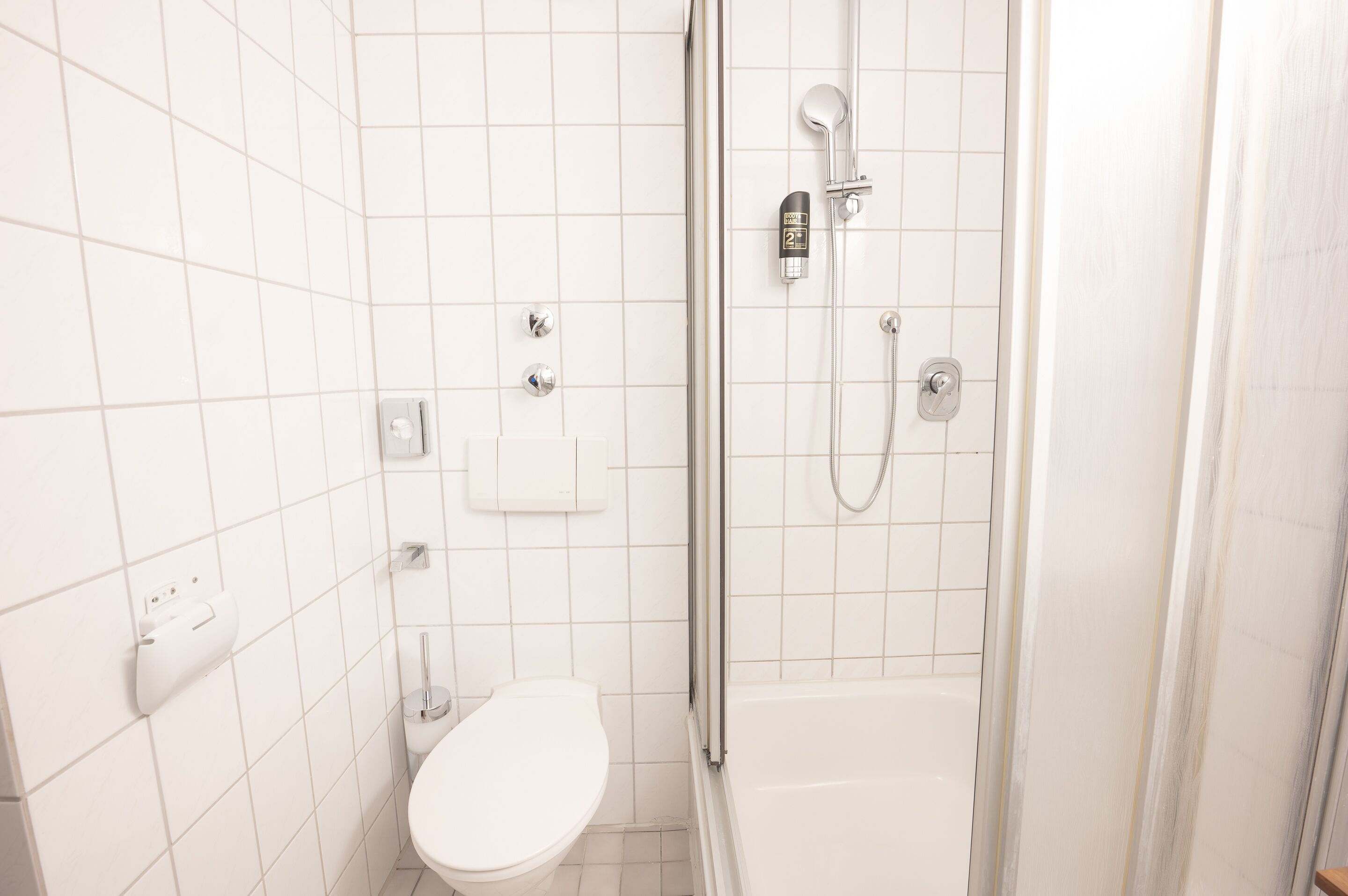 A modern bathroom with white tiles, a toilet, a bathtub, and a shower with a handheld showerhead.