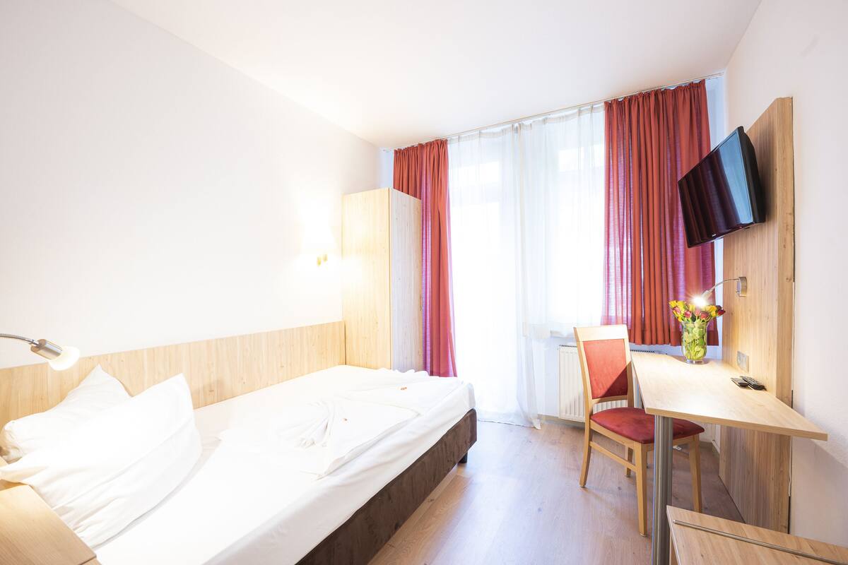 A hotel room with a bed, a desk, a chair, a TV mounted on the wall, and a window with curtains.