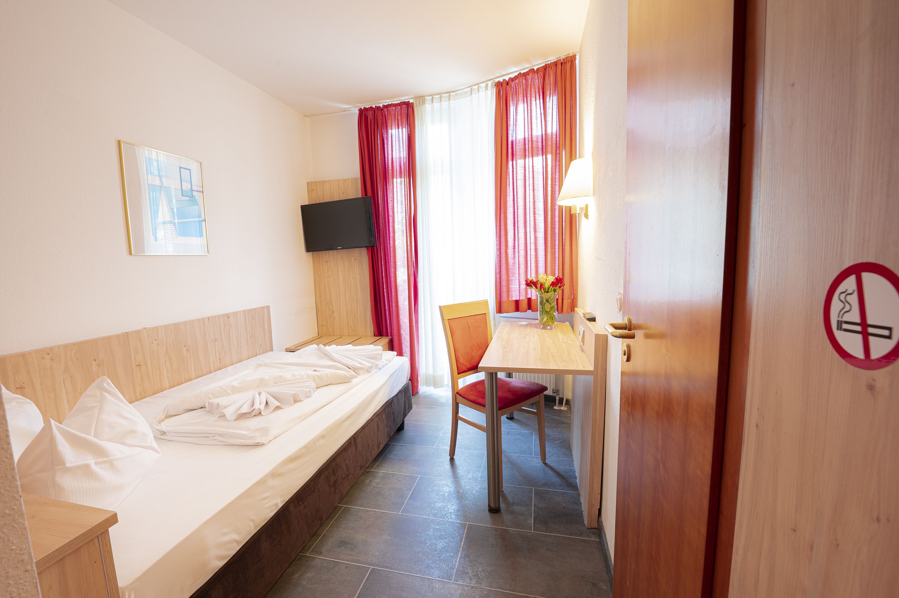 A hotel room with a bed, a desk, a chair, a TV mounted on the wall, and a door with a no-smoking sign.