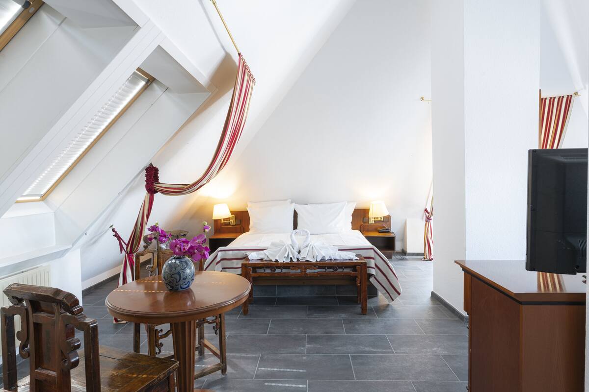 A hotel room with a bed, bedside tables, a TV, a wooden chair, a round table with a vase, and a view of the sky through a skylight.