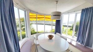 A dining area with a round table, white chairs, and a bowl. There are large windows with curtains, a view of greenery and water outside.