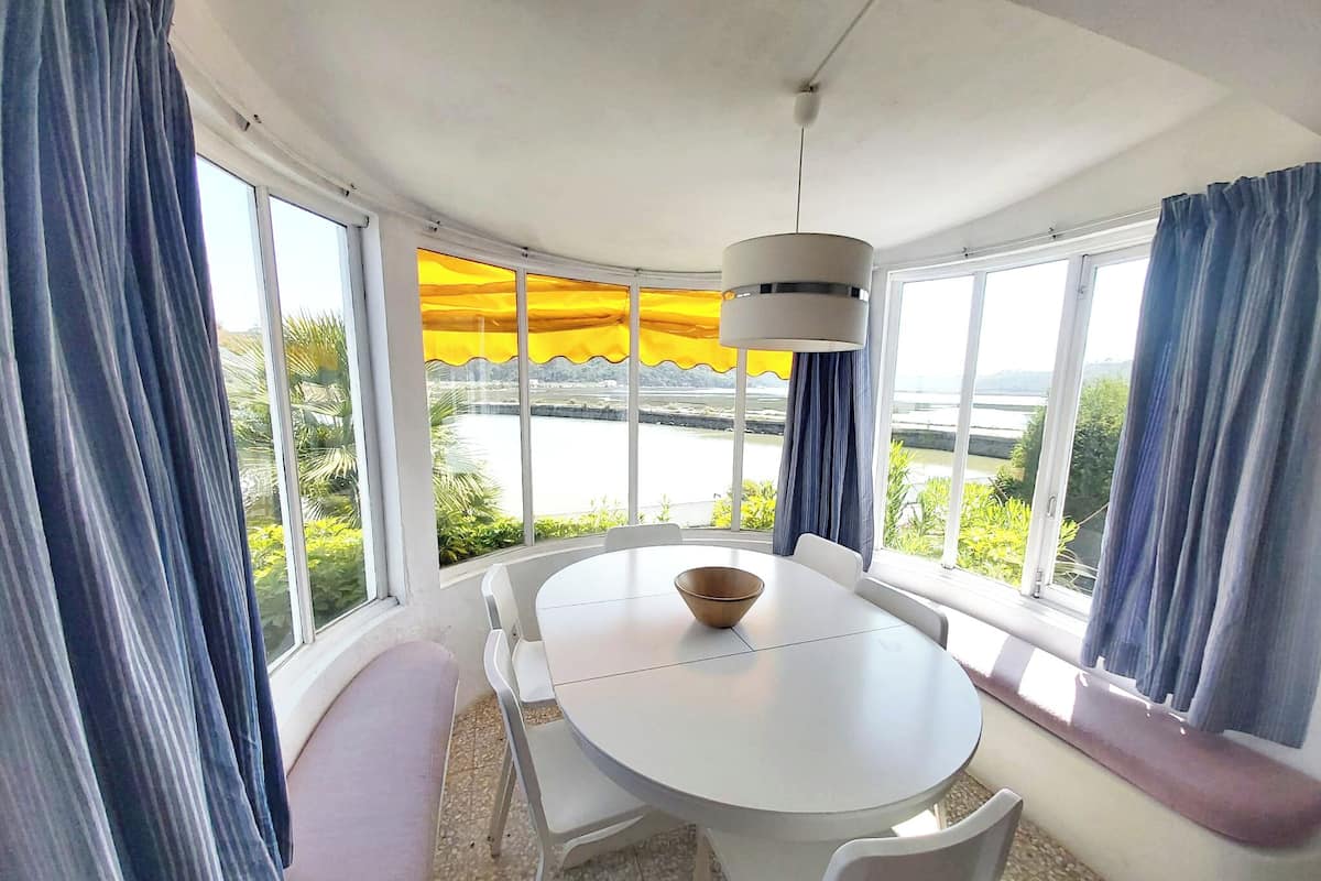 A dining area with a round table, white chairs, and a bowl. There are large windows with curtains, a view of greenery and water outside.