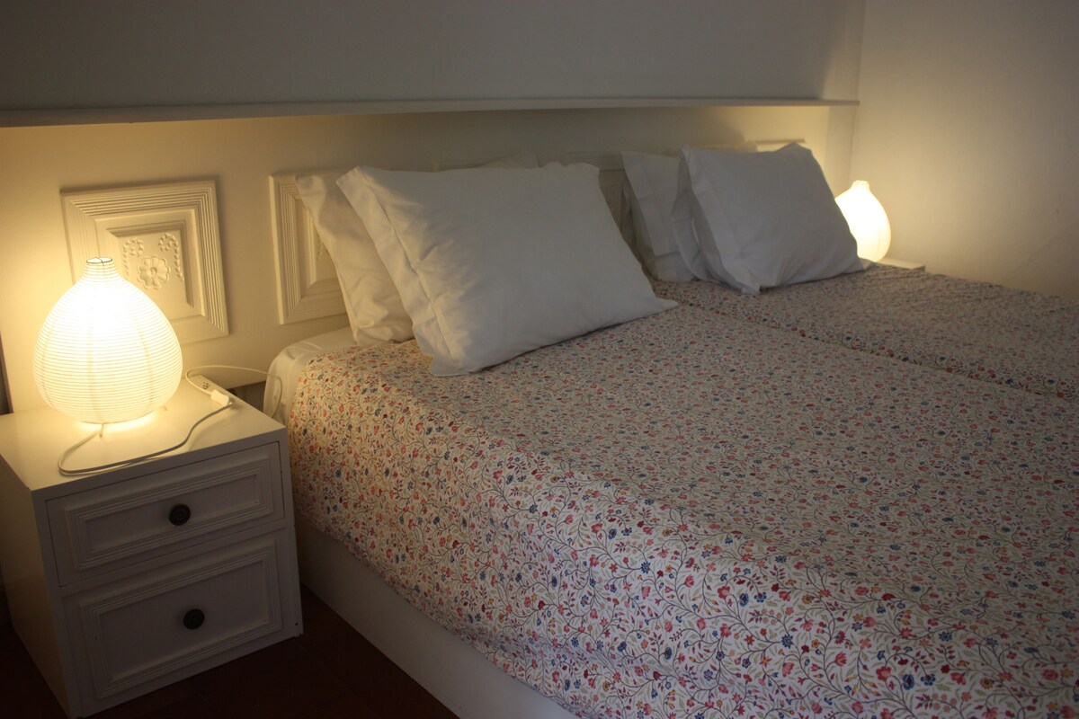 Una cama bien hecha con ropa de cama floral, una lámpara de noche y una mesita de noche con cajones.