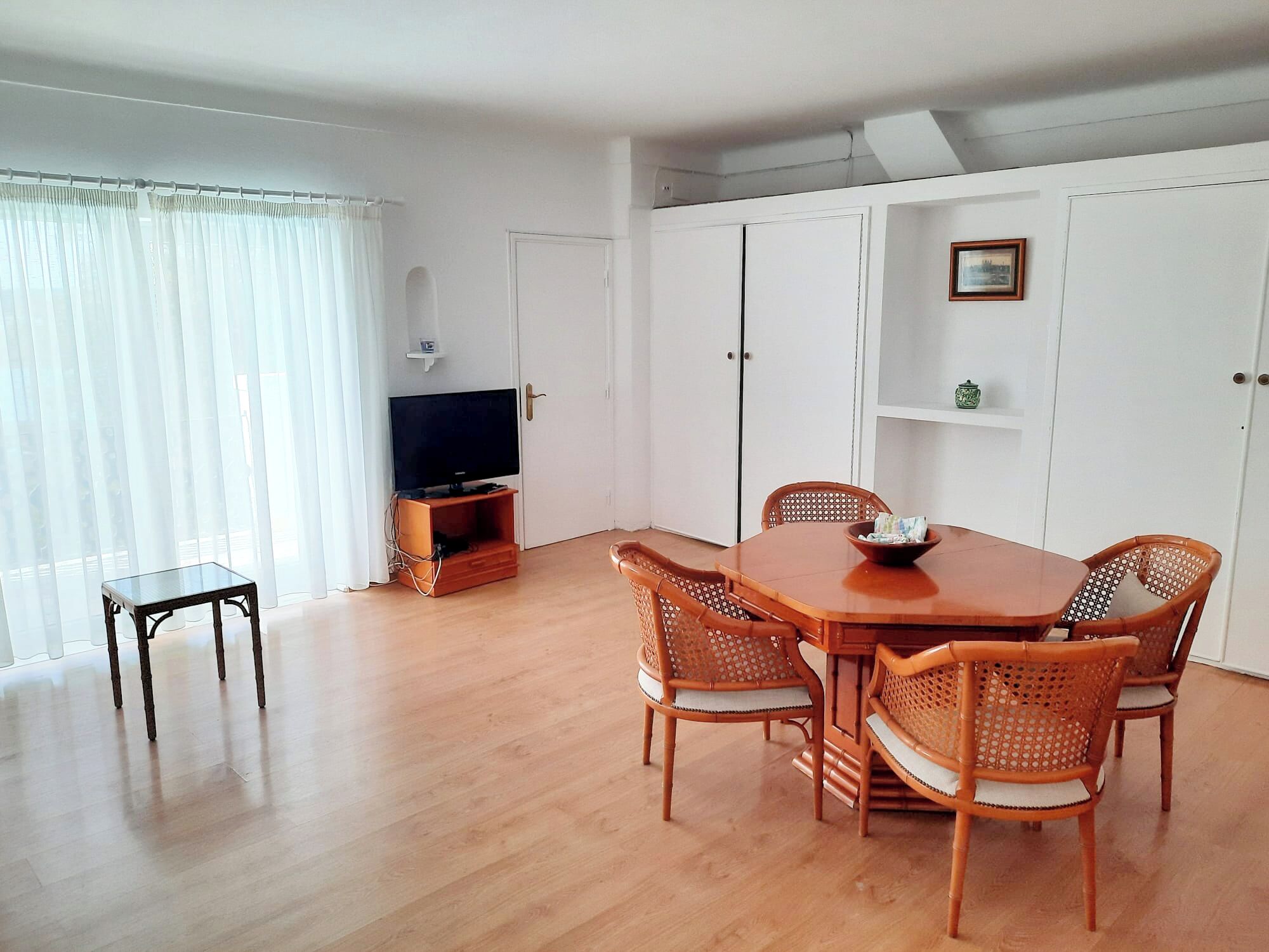 A dining area with a wooden table and chairs, a television on a stand, and white walls.