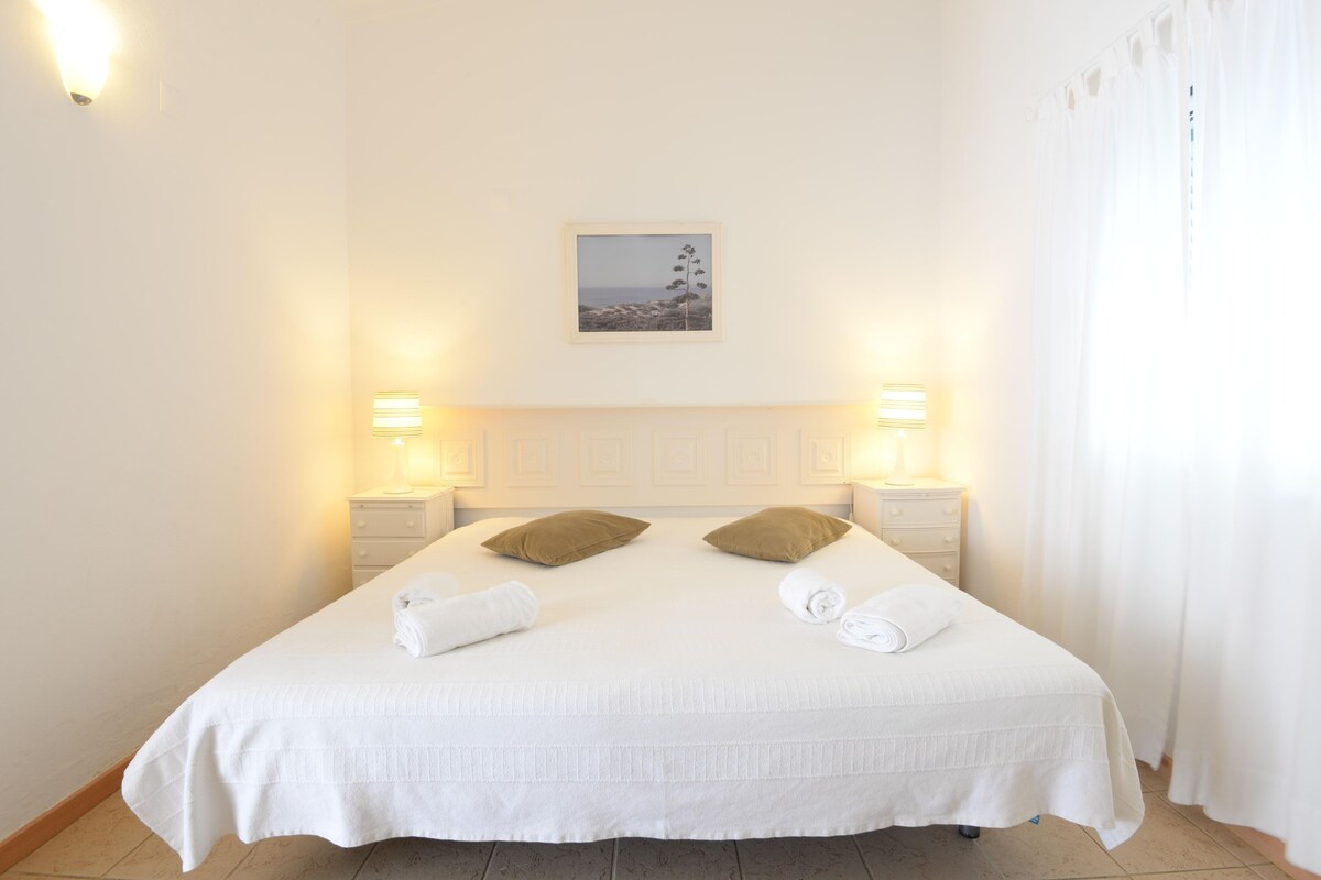 A neatly made bed with white linens and two brown pillows, flanked by two bedside tables with lamps, in a room with a plain white wall and a framed picture above the bed.