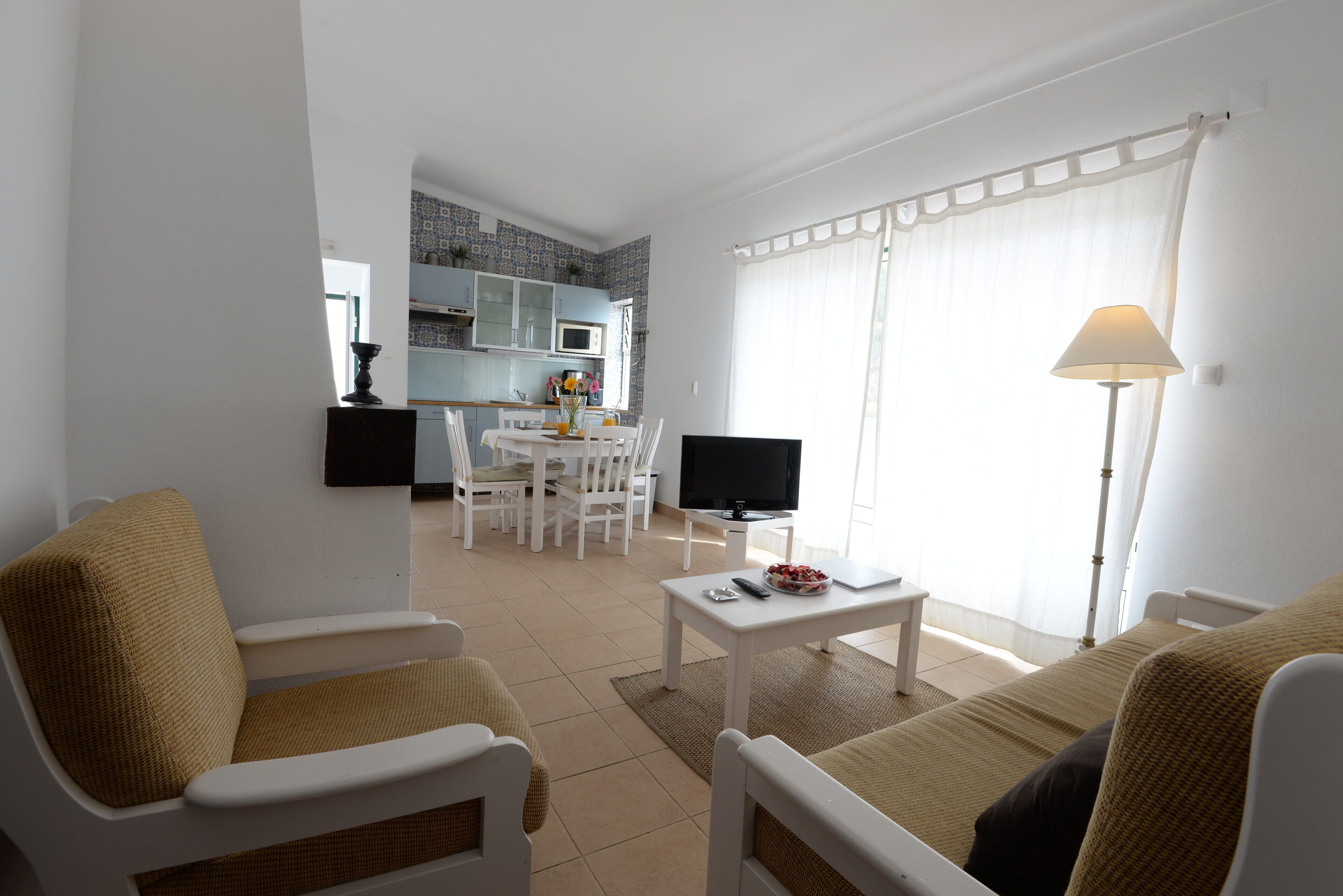 Un salon avec un téléviseur, un canapé et des fauteuils, un coin repas avec une table et des chaises, et une cuisine à l’arrière-plan.