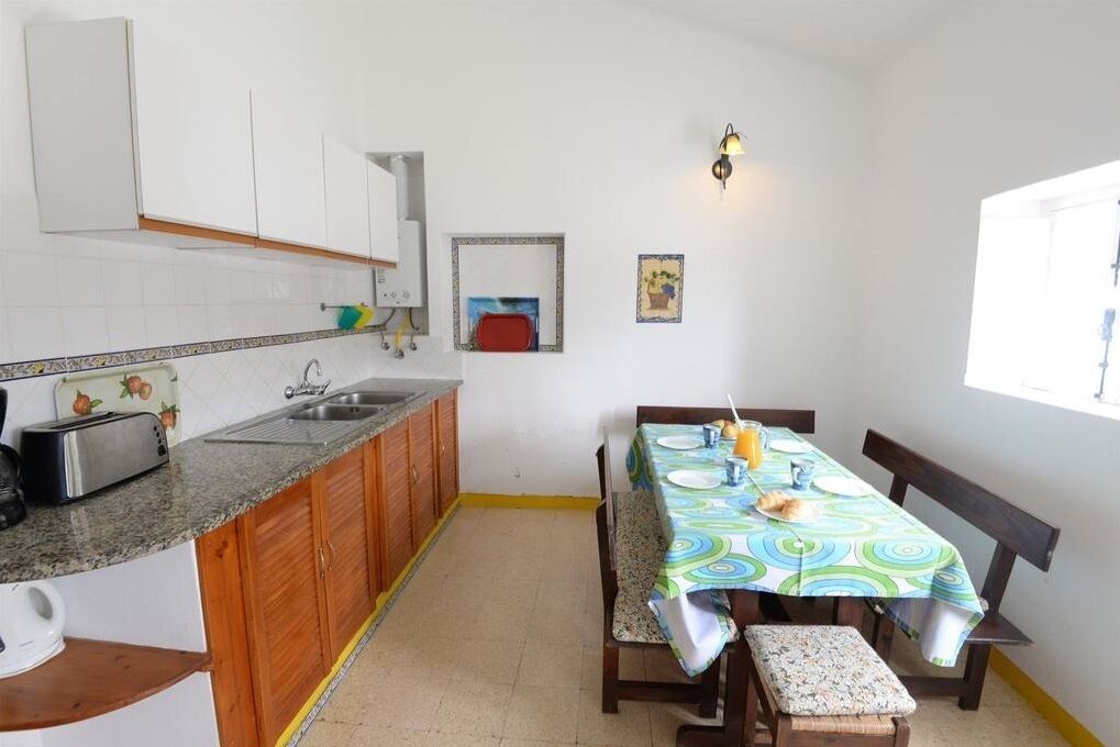 A compact kitchen with wooden cabinets, a granite countertop, a dining table with a colorful tablecloth, and a bench.