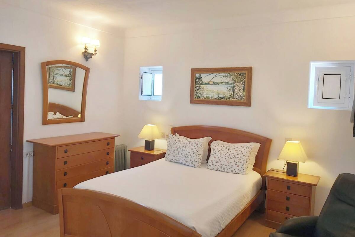 A bedroom with a wooden bed, two bedside tables with lamps, a dresser, and two wall-mounted picture frames.