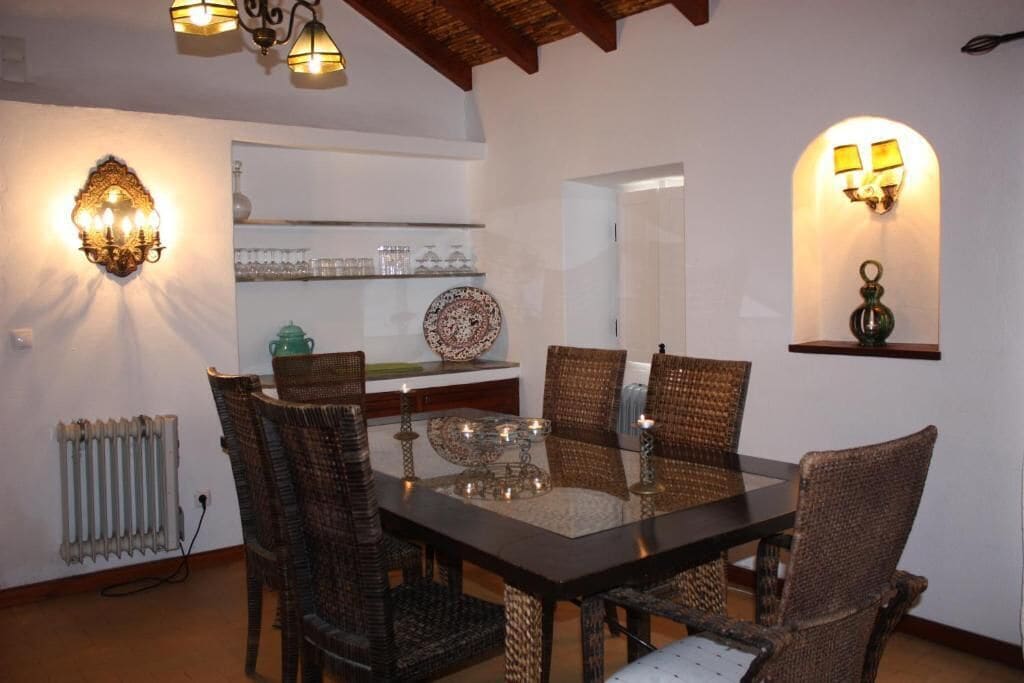 A dining area with a glass table surrounded by wicker chairs, wall-mounted sconces, and a shelf with glassware.