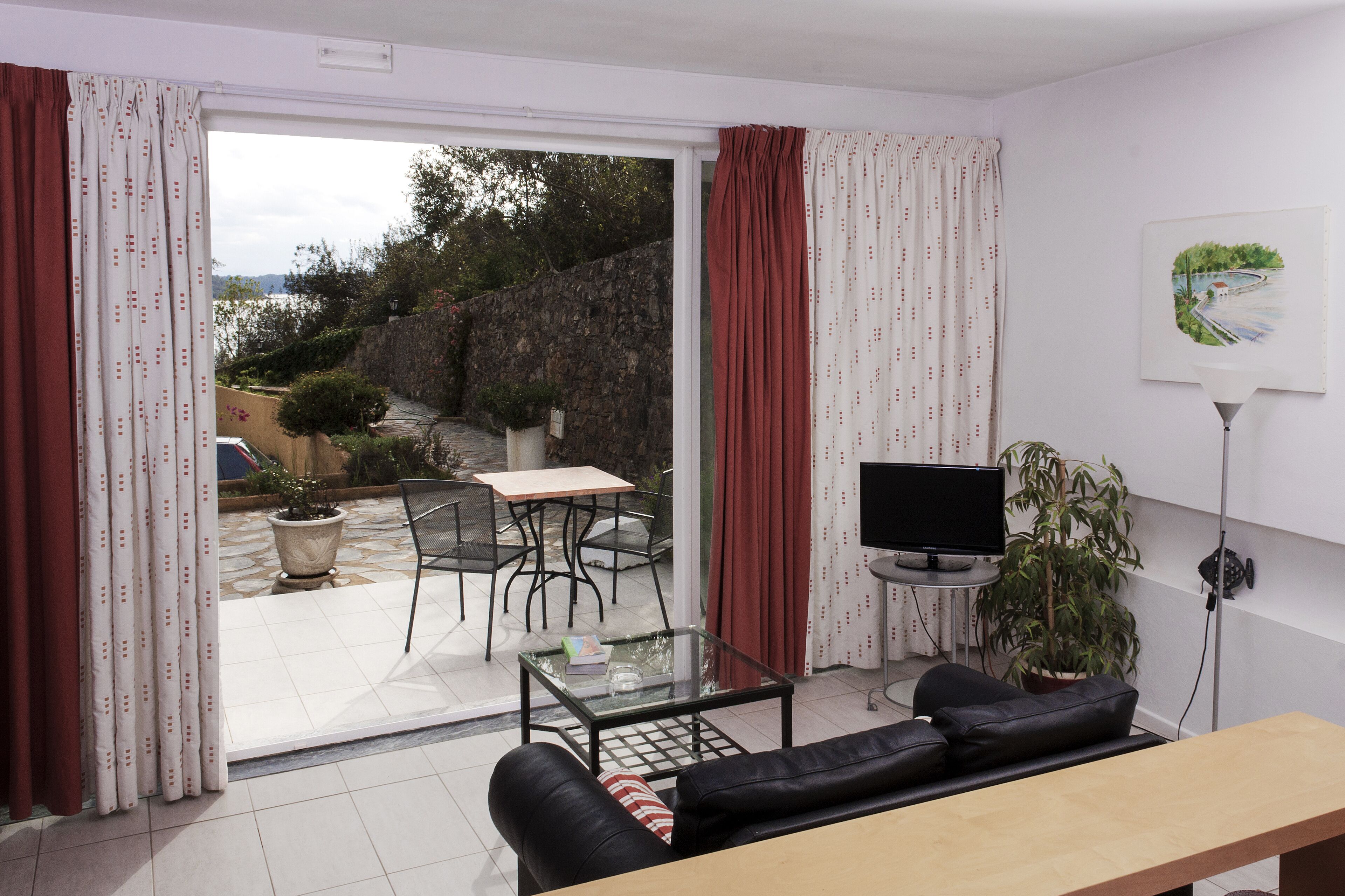 Una sala de estar con sofá, una mesa de centro, un televisor sobre un mueble bajo, y vistas a un patio exterior con mesa y sillas.