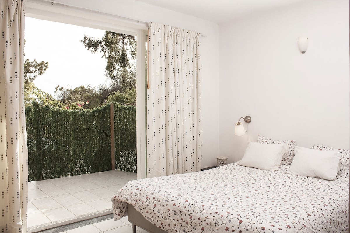 A bedroom with a bed, a nightstand, a lamp, and a view of a garden through a sliding door.