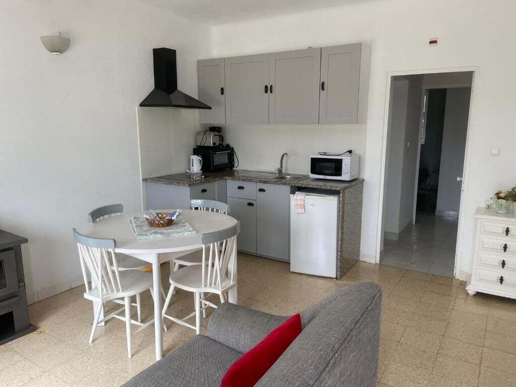 A compact kitchen with white cabinets, a microwave, and a coffee maker. There is a dining table with chairs and a grey sofa with a red cushion.