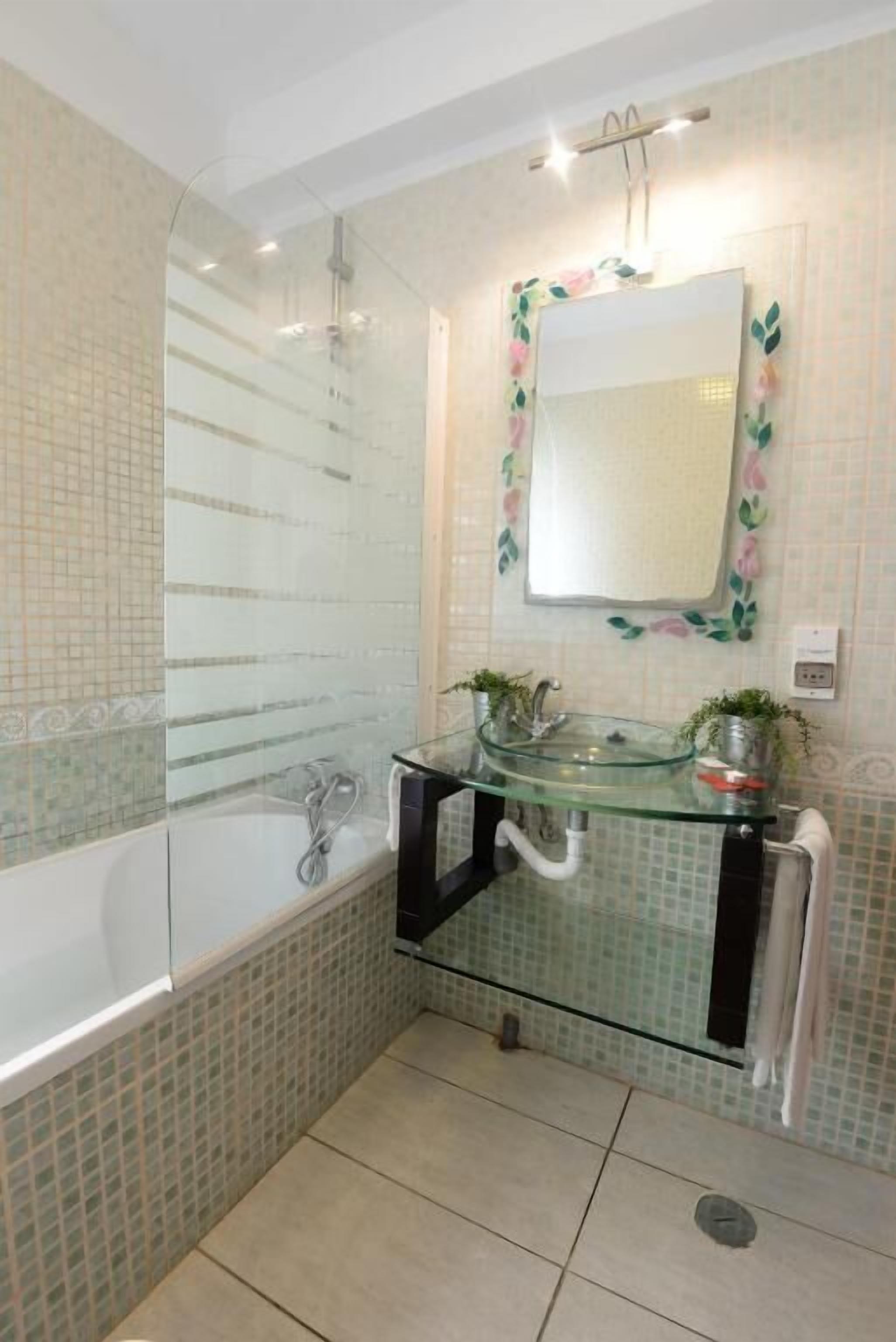 A bathroom with a glass-enclosed shower, a glass sink with two plants, and a mirror with floral decorations.