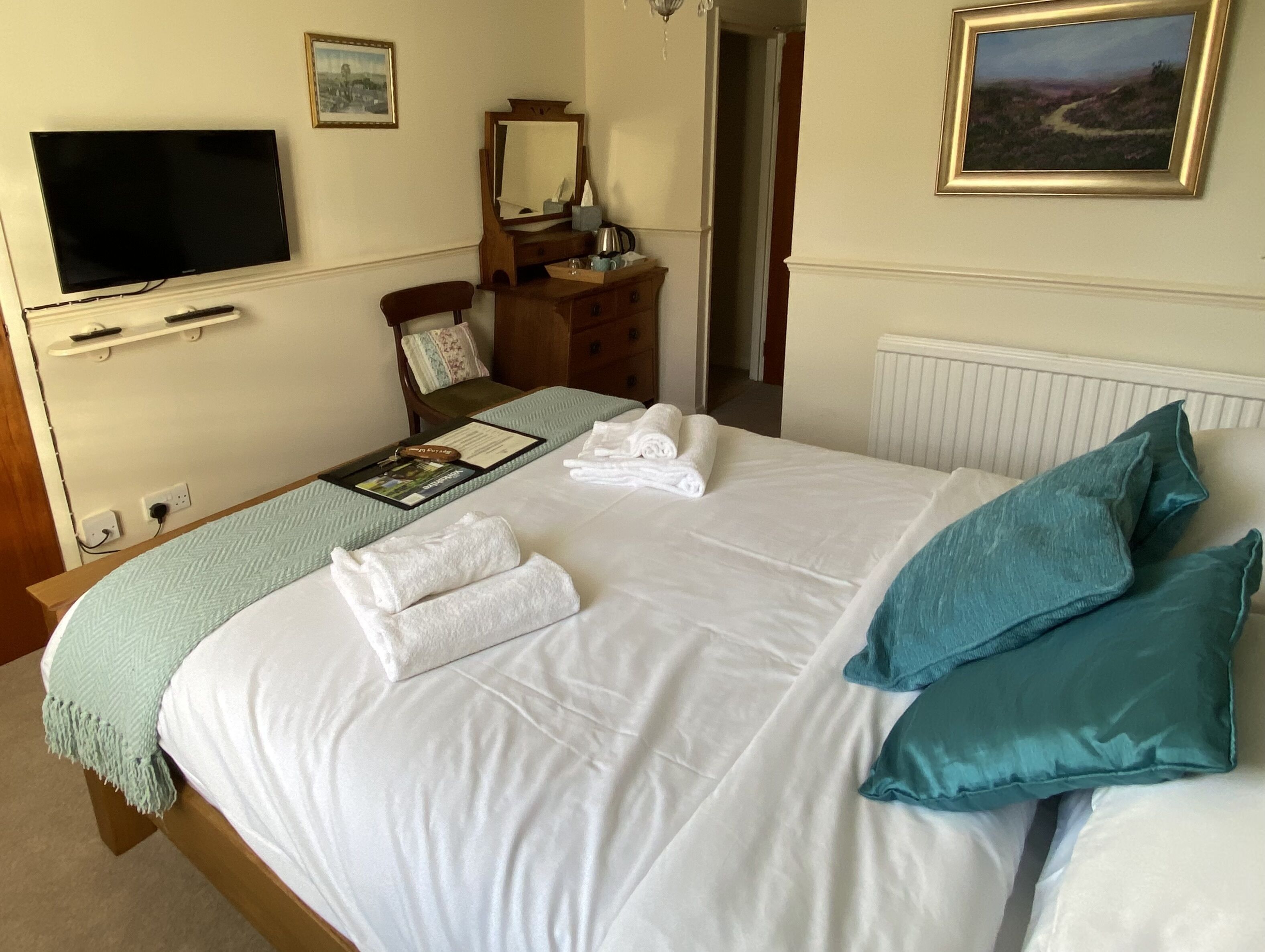 A neatly made bed with white linens and teal pillows, a flat-screen TV mounted on the wall, a wooden dresser, and a framed landscape painting.