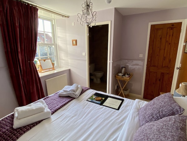 A hotel room with a bed, a window with curtains, a chandelier, a wooden door, and a small table with toiletries. - Plumpton Court (York)