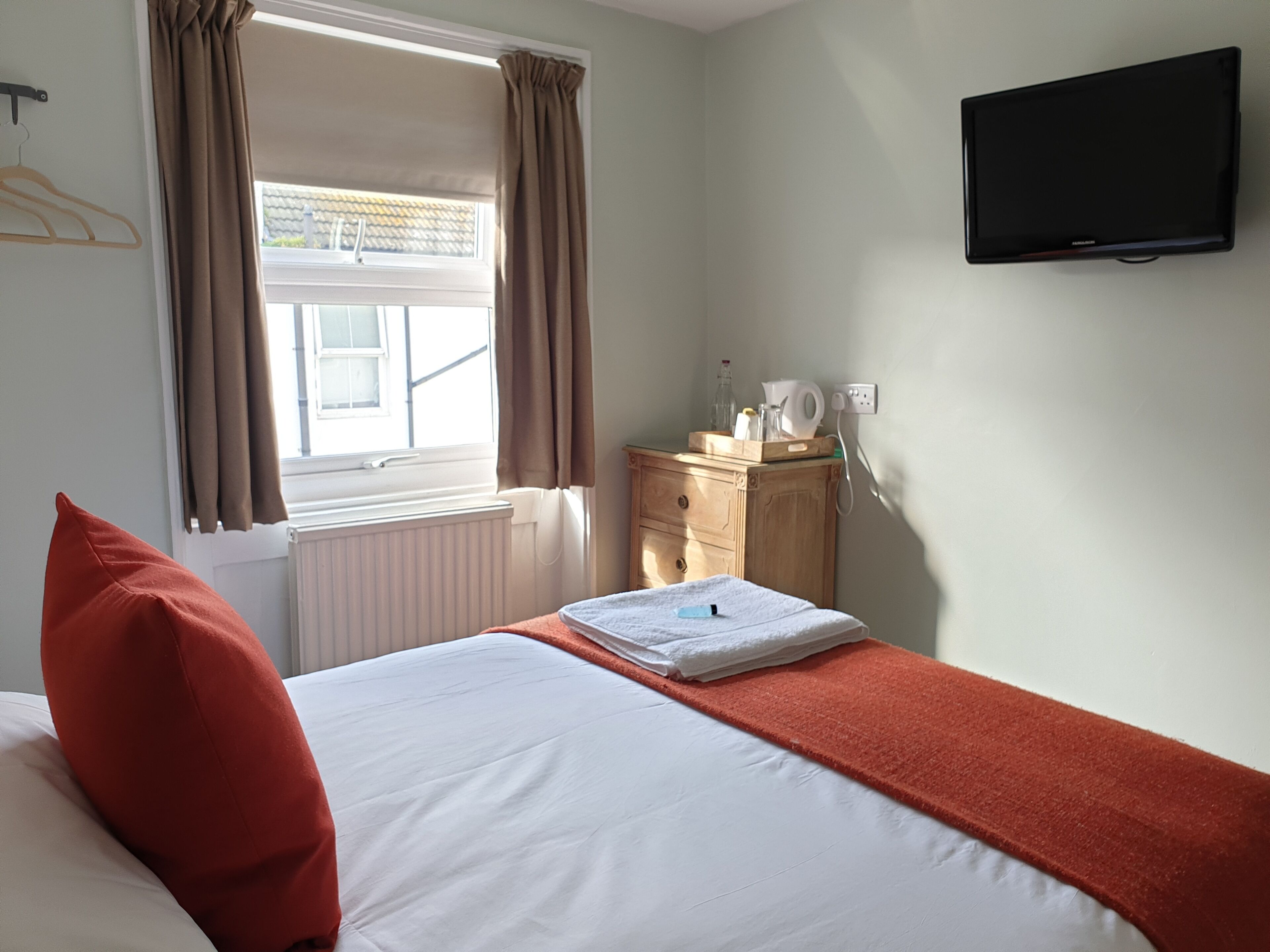 A hotel room with a bed, a TV mounted on the wall, a small wooden dresser, and a window with curtains.