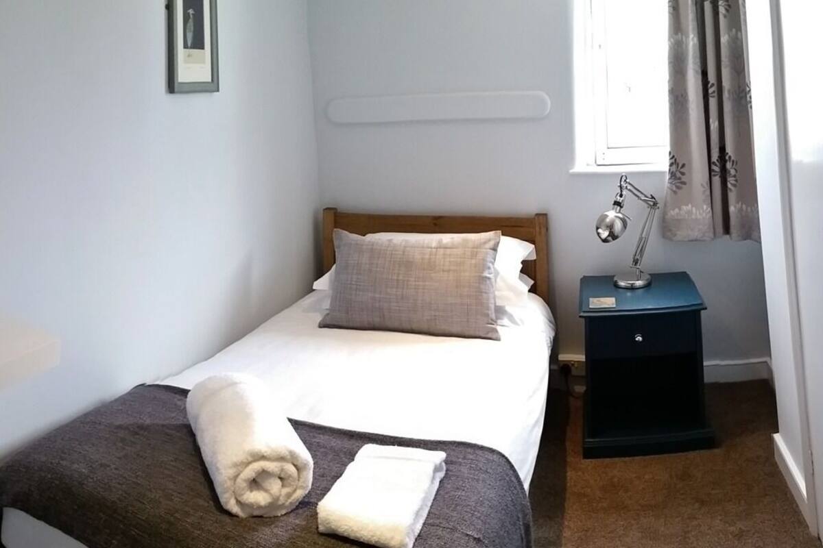 Un lit bien fait, recouvert d’une couverture blanche et marron, avec une lampe de chevet et une petite table de nuit.