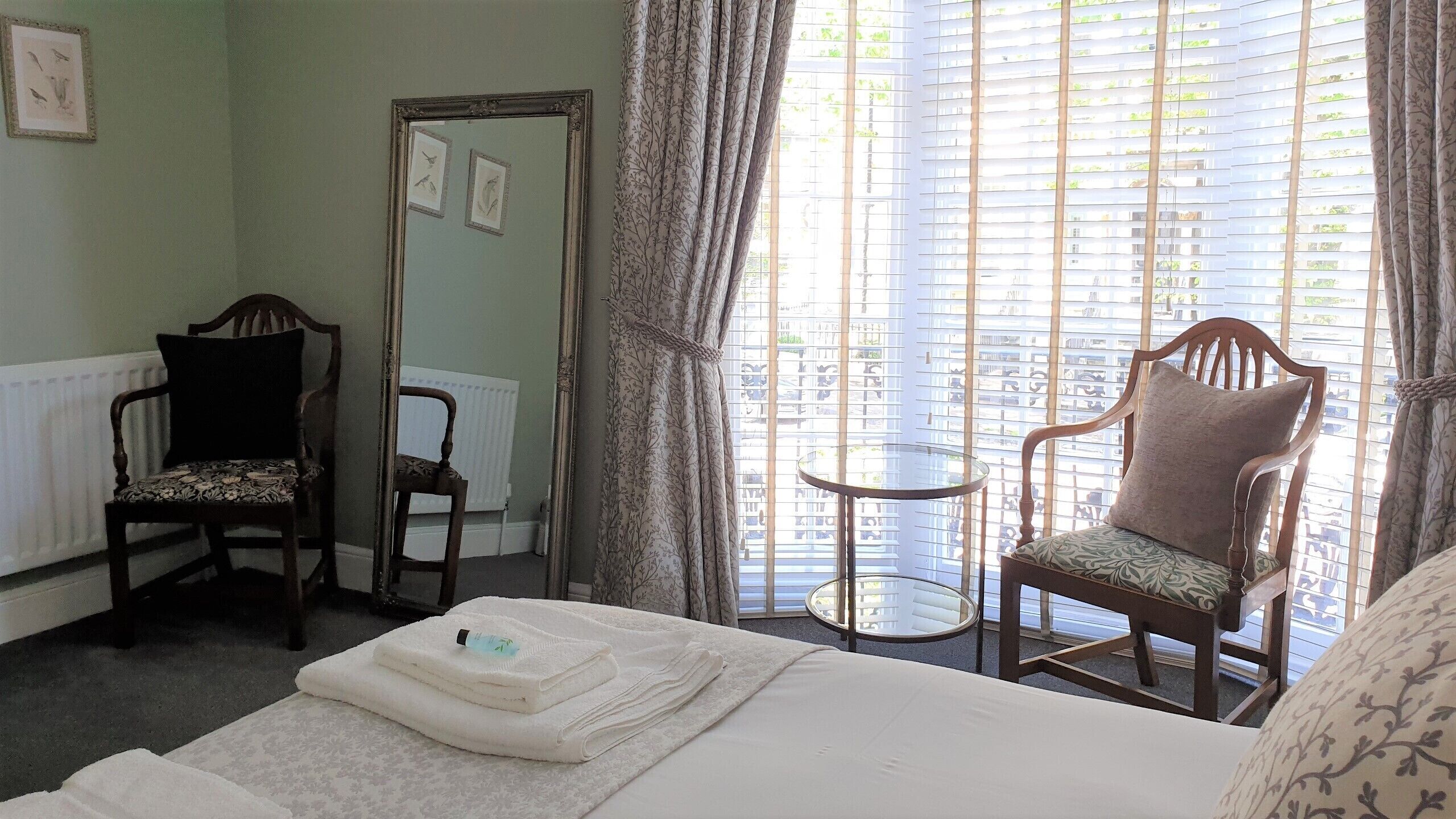 A hotel room with a bed, two chairs, a small table, and a window with blinds.