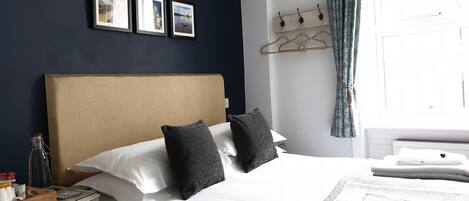 A neatly made bed with white linens and two dark pillows, a wooden nightstand with a bottle and a tray, and a wall with three framed pictures.