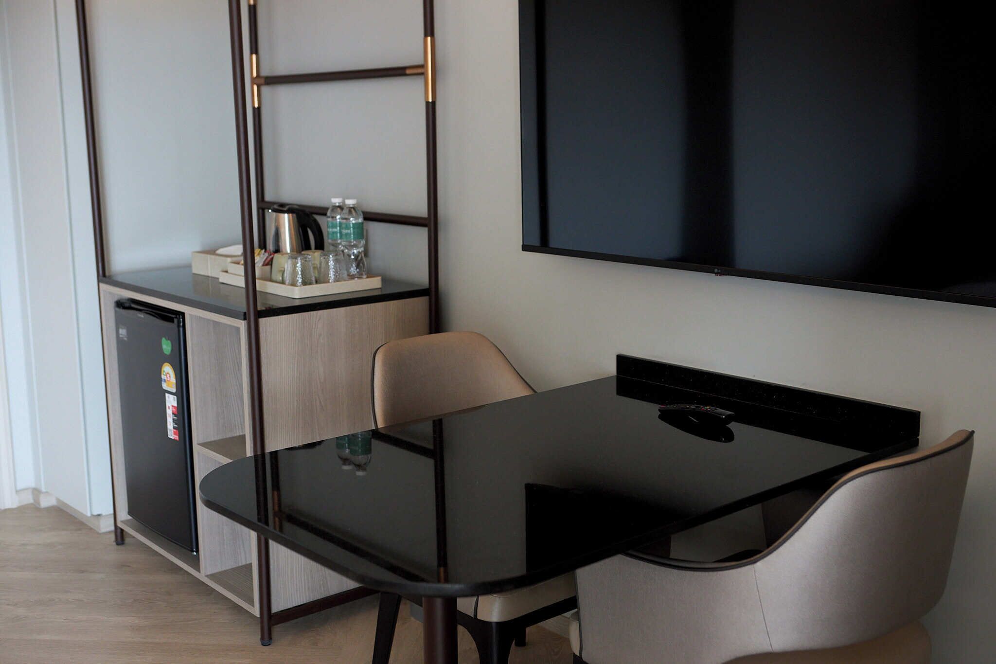 A modern room with a glass-top table, a chair, a small kitchenette, and a flat-screen TV mounted on the wall.
