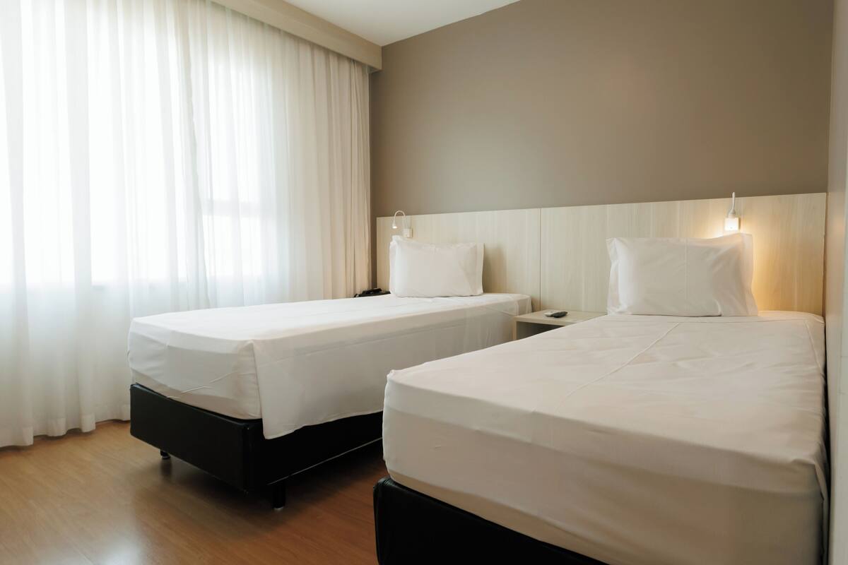 Dos camas individuales con sábanas blancas, suelo de madera y una ventana con cortinas traslúcidas.