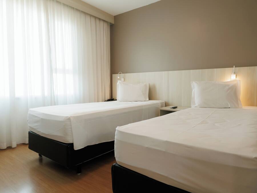 Two single beds with white linens, a wooden floor, and a window with sheer curtains.