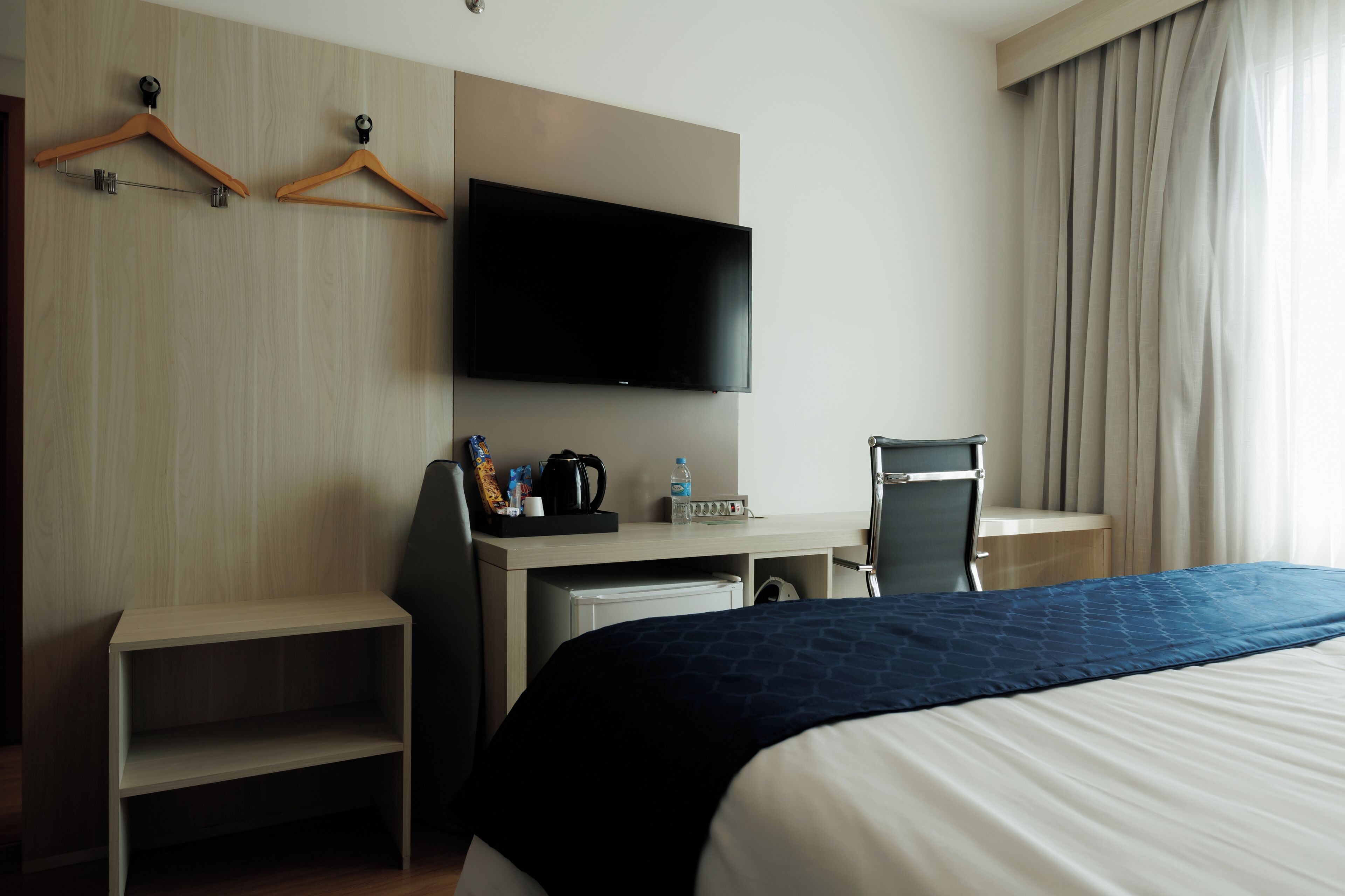 Una habitación de hotel con cama, escritorio con televisor de pantalla plana, silla, cafetera y estante.