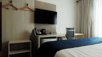Una habitación de hotel con cama, escritorio con televisor de pantalla plana, silla, cafetera y estante.